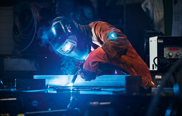 Soudeur avec casque de soudage et tenue de protection effectuant un soudage MAG de pièces métalliques dans un atelier