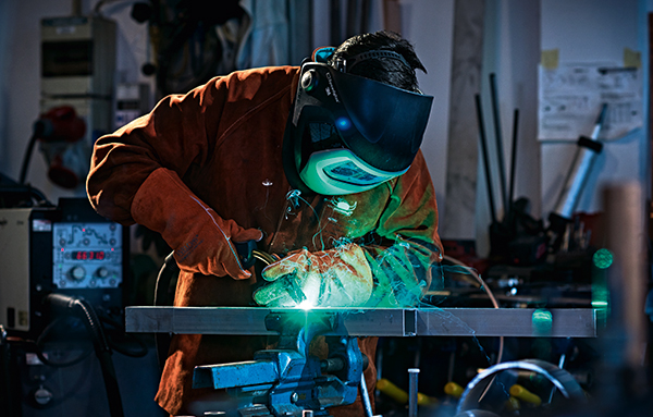 Soudeur avec casque de soudage et tenue de protection effectuant un soudage MIG de pièces métalliques dans un atelier