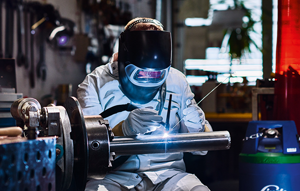 Soudeur avec casque de soudage et tenue de protection effectuant un soudage TIG de pièces métalliques dans un atelier