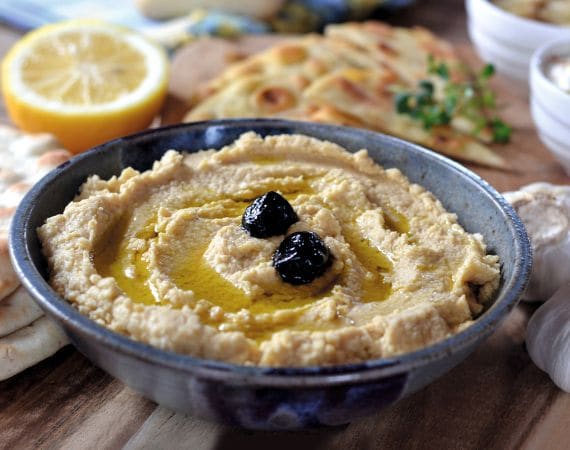 Schüssel mit Hummus. Darauf 3 schwarze Oliven als Deko drapiert. Außenrum eine halbe Zitrone sowie ein Fladenbrot