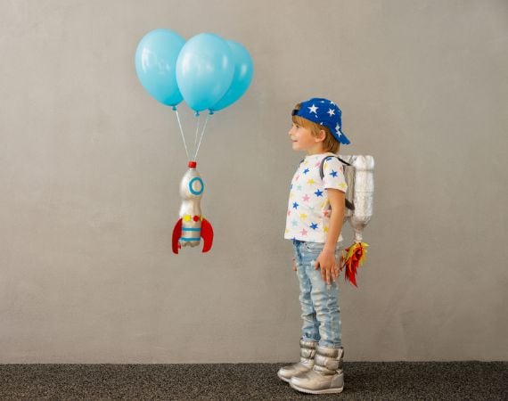 kleiner Kunde mit Kappe und Rucksack schaut auf 3 schwebende Ballons an denen eine mit Alufolie ummantelte PET-Flaschen hängt, die eine Weltraumrakete simulieren soll